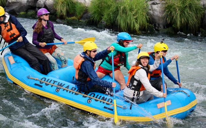 a group of students wearing helmets and life jackets paddle a blue raft through whitewater